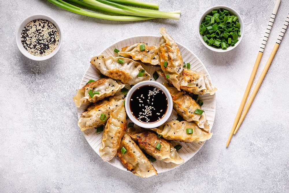 Stekta gyoza dumplings på tallrik med sojasås, salladslök och ätpinnar, fotograferade ovanifrån.