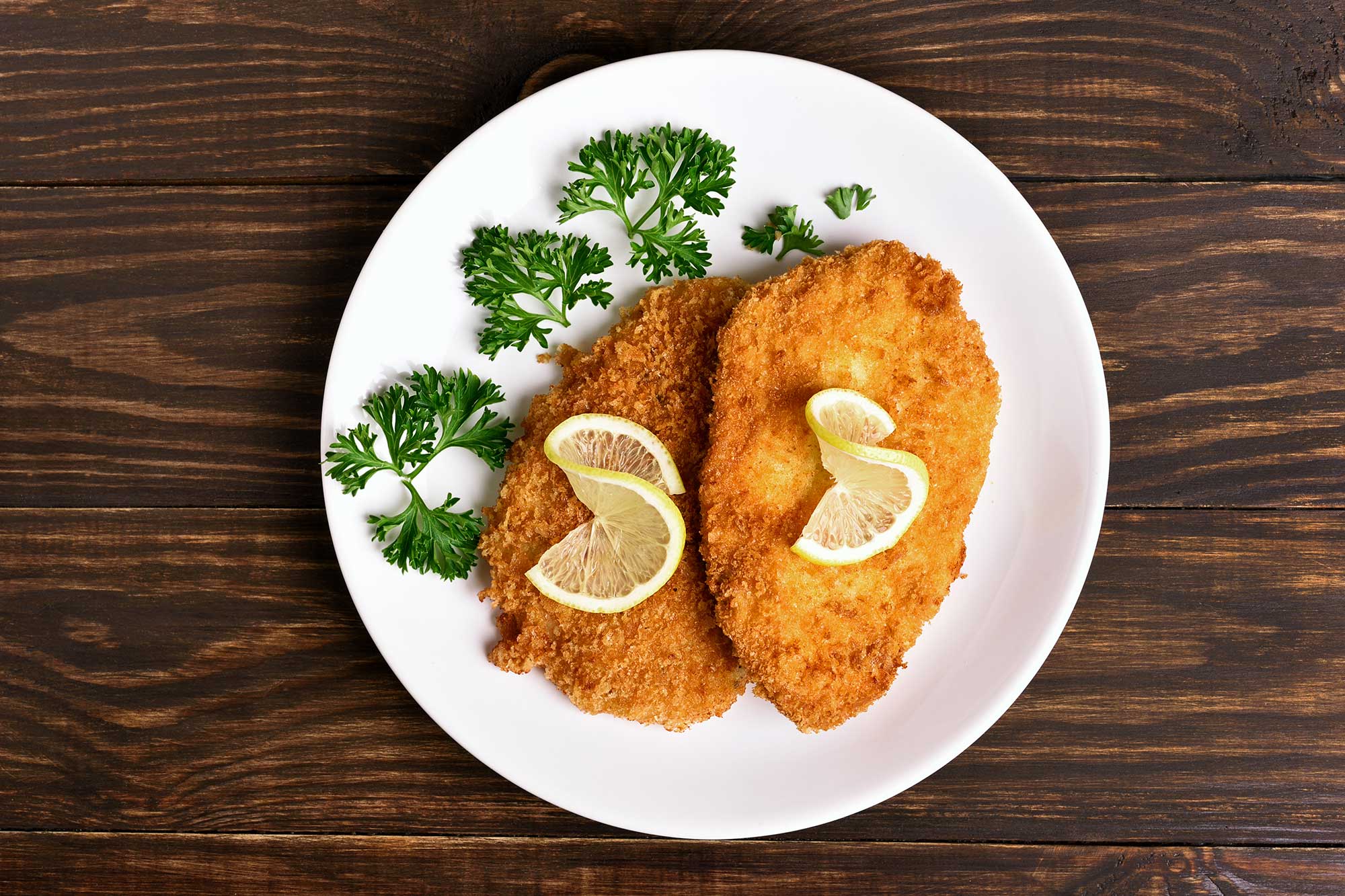 Två gyllene kycklingschnitzlar på vit tallrik med citronskivor och persilja mot träbakgrund.